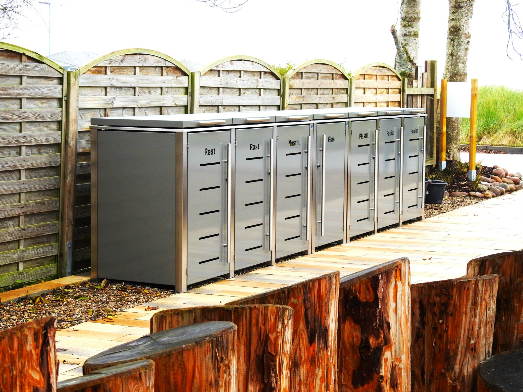 Mehrere Mülltonnenboxen in moderner Gartenumgebung mit Beschriftung Papier, Glas, Plastik und Rest vor Holzsichtschutz
